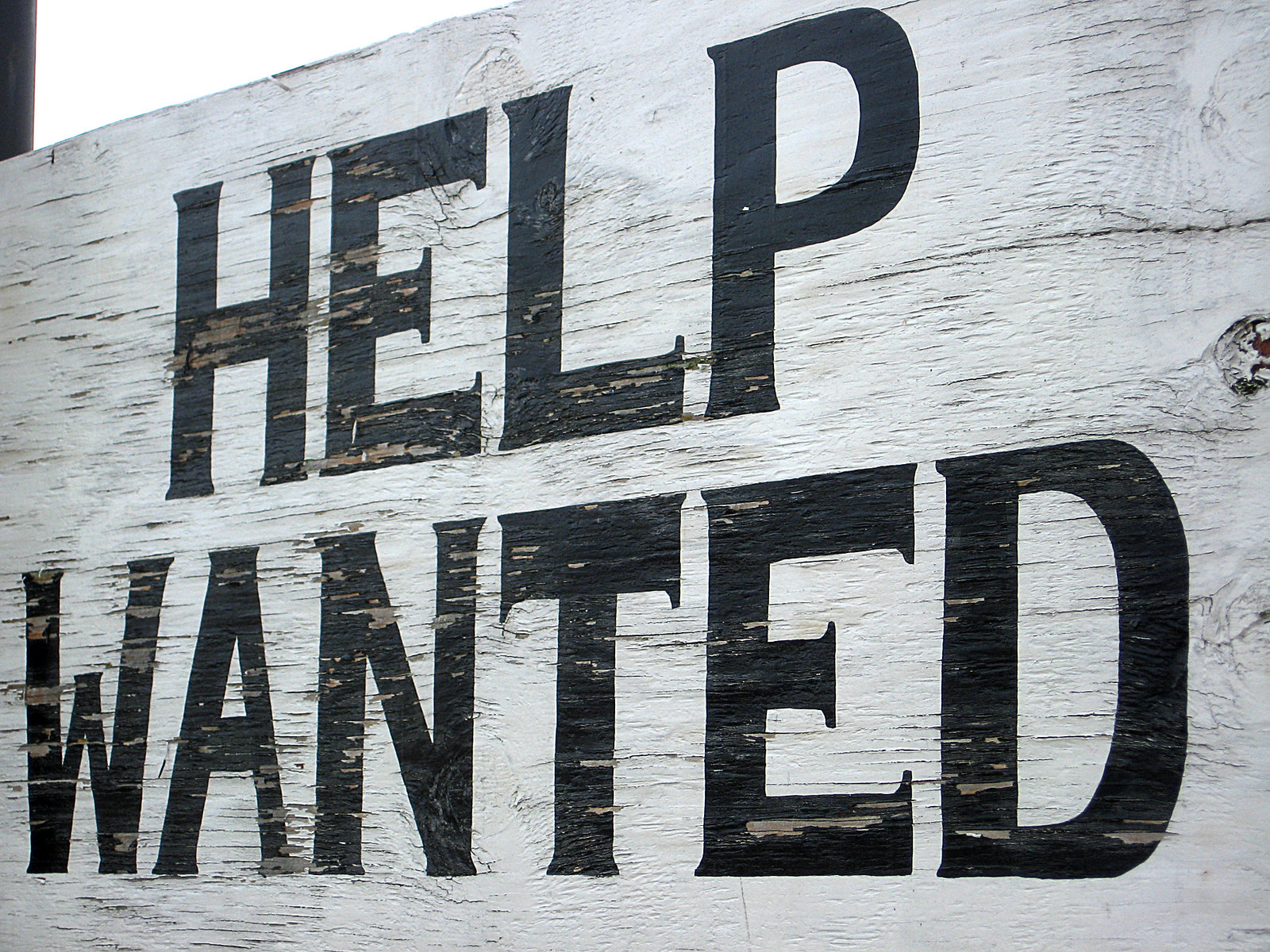 Photograph of a sign displaying the text "help wanted". Black letters painted on a white wooden background.